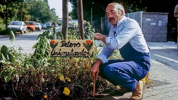 Jüngerer Peter Lustig in den Achtzigern vor einem kleinen Unkraut-Garten in der Stadt