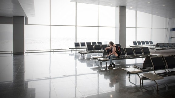 Leere Sitze am Gate eines Flughafens, einzelene junge Frau sitzt mit Rucksack auf Sitz und schaut zur Seite.