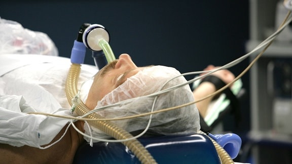 Patient wird künstlich beatmet: Blick auf liegenden Patienten, seitlich links oben. Patient ist an einen Schlauch sowie kleinere Schläuche oder Kabel angeschlossen.