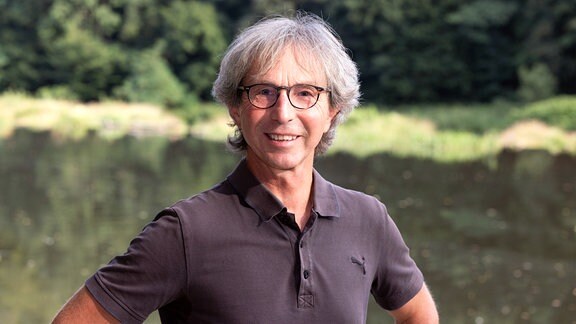 Portait von Mann mit halblangen graumelierten Haaren, schwarzer Brille und dunklem kurzärmligen Polo-Shirt, lächelnd