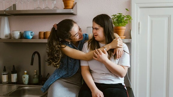 Zwei junge Frauen mit braunen, längeren Haaren, eine mit Down-Syndrom, lächeln sich an und umarmen und berühren sich in einer Küche einer Altbauwohnung
