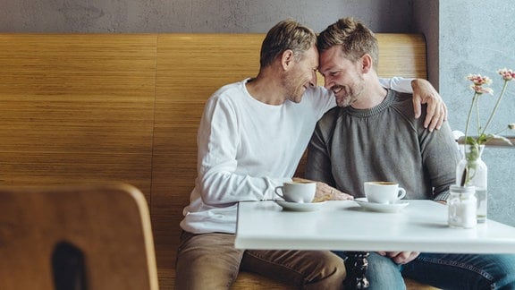 Zwei Männer mit Drei-Tage-Bart sitzen in Kaffee, glücklich lachend, Stirn an Stirn, Hand-Berührung und Arm um Schulter
