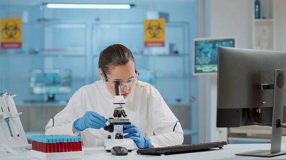 Eine Frau mit weißem Kittel und Schutzbrille sitzt an einem Mikroskop in einem Labor.