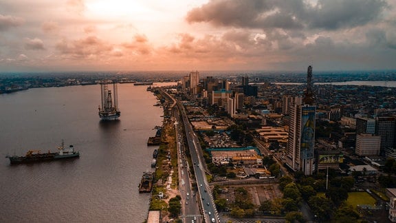 Luftaufnahme mit Blick auf Hafen am Fluss, mehrspurige Straße, Hochhäuser in Lagos, tiefstehende Sonne, warme, rötliche Farben