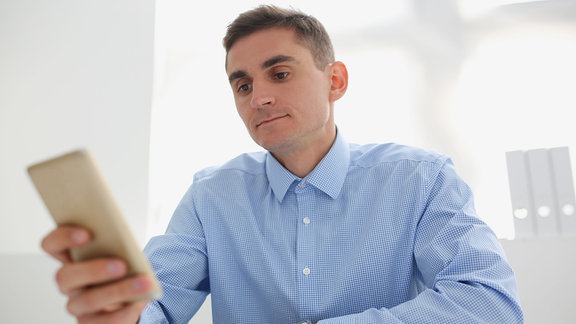 Junger Mann im blauen Hemd schaut auf den Smartphone-Bildschirm, während er an seinem Schreibtisch sitzt.