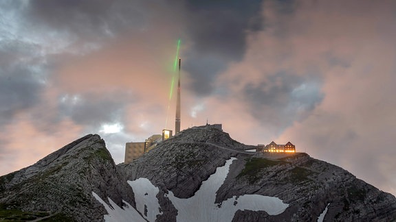 Ein grüner Laserstrahl geht auf eine Turmspitze. Dunkle Wolken im Hintergrund.