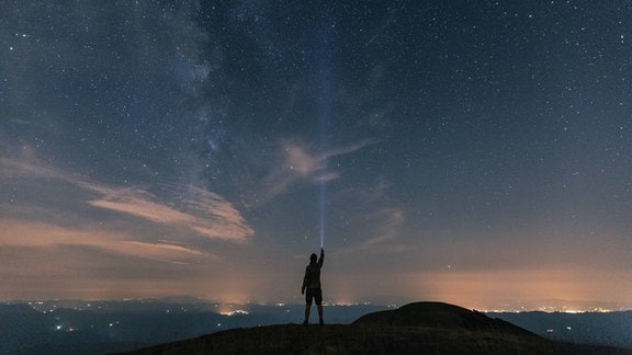 Symbolfoto: Silhouette eines Menschen leuchtet mit einer Taschenlampe in den Sternenhimmel.