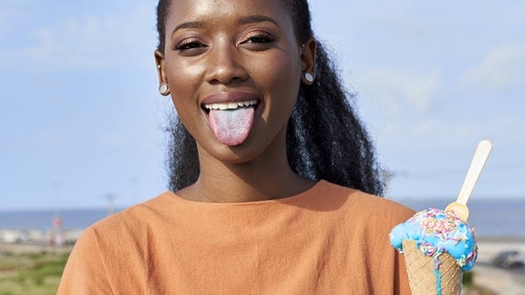 Junge Frau lacht bei sonnigem Wetter, streckt leich bläulich gefärbte Zunge raus und hält Eistüte mit blauem Eis in der Hand, das leicht tropft