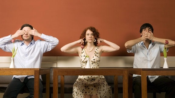 Drei junge Menschen, Mann, Frau, Mann, sitzen nebeneinander in Café oder Restaurant, links hält Augen zu, Mitte die Ohren und rechts den Mund