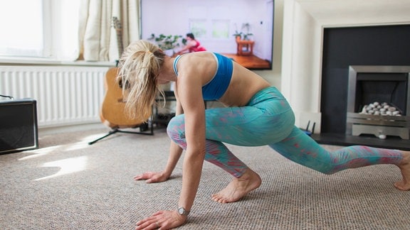 Frau mit blonden Haaren mit Pferdeschwanz und Sportkleidung in Sprinter-Start-Pose im Wohnzimmer blickt zum Fernseher, wo ein Yoga-Kurs läuft