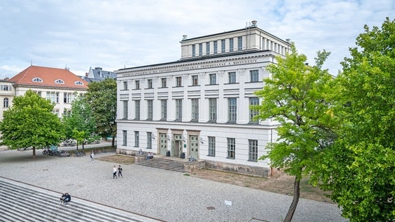 Blick auf ein weißes Gebäude, neben dem grüne Bäume stehen. Der Vorplatz ist hellgrau gepflastert.