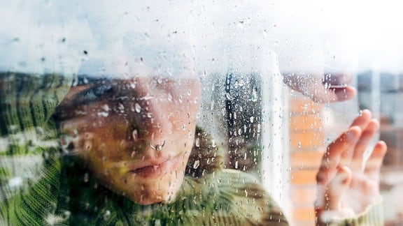 Junge Frau mit Pullover schaut mit Stirn auf Unterarm melancholisch durch ein verregnetes Fenster. Ansicht von außen, viel Unschärf.