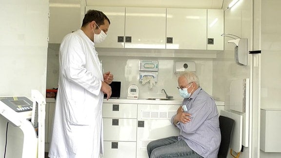 Behandlungszimmer, Seitenansicht: Arzt mit Kittel und Mund-Nase-Bedeckung steht vor sitzendem älteren Patienten mit Mund-Nase-Bedeckung. Im Hintergrund Schrank, Waschbecken.