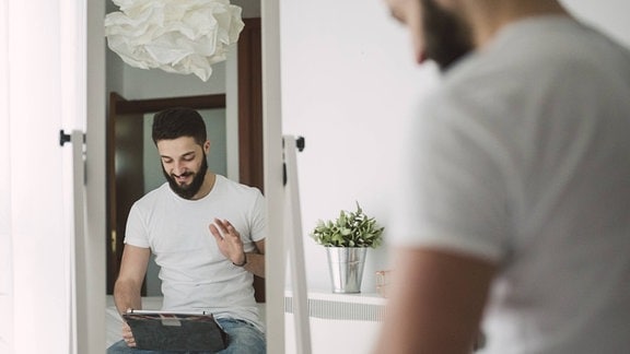 Mann mit dunklen Haaren und Bart sitzt vor einem Spiegel und Blickt auf Tablet-Computer mit erhobener Hand. Heller Raum.