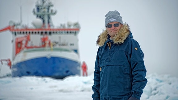 Das Bild zeigt einen warm angezogenen Mann mit Mütze und blauem Winteranorak mit Fellbesatz. Im Hintergrund der Bug eines Schiffes mit blauem Rumpf und weißem Aufbau.