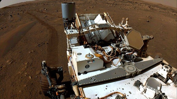 Der NASA-Rover Perseverance auf der Marsoberfläche. Die Kamera des Rovers blickt von oben auf ihn runter und zeigt seinen Vorbau, der mit wissenschaftlichen Geräten bestickt ist. Im Hintergrund erkennt man den braun-rötlichen Marsboden und seinen bräunlichen Himmel.