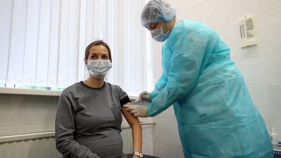Eine schwangere Frau in einem grauen Kleid trägt einen Mundschutz und sizt in einem Behandlungszimmer. Sie bekommt eine Impfung von einer Person in Coronaschutzkleidung.