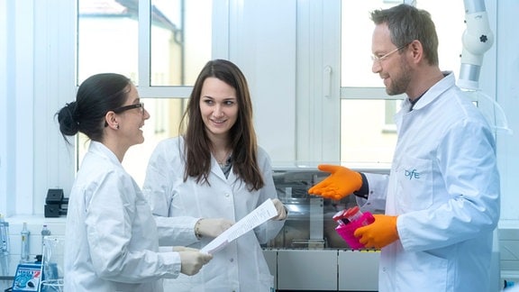 Drei Wissenschaftler mit weißen Kitteln, links zwei Frauen, rechts ein Mann, stehen in einem Labor.