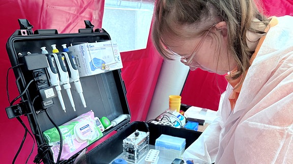 Eine Forscherin in Schutzkleidung sortiert Laborgeräte in einem schwarzen Hartplastik-Koffer. Innerhalb des Koffers sieht man Gummihandschue, Pipetten, eine Zentrifuge und andere Laborgeräte.