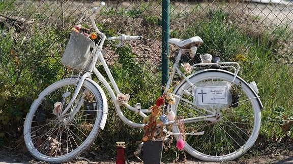 Weiß gestrichenes Fahrrad mit Blumen und Todesanzeige am Rahmen lehnt an einem Zaun.
