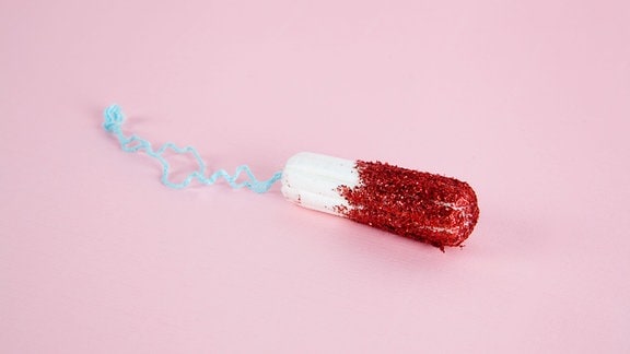 Ein Tampon mit rotem Glitzer anstatt Blut liegt auf einem rosa Untergrund.