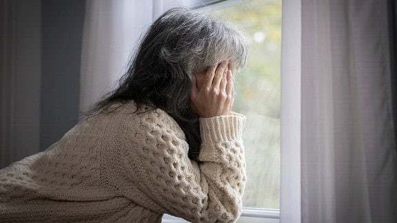 Ältere weiße Frau am Fenster, die Hände vor dem Gesicht, sieht deprimiert aus