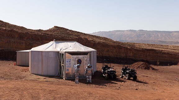 Der Blick auf das Habitat der sechs analogen Astronauten. Draußen stehen zwei von ihnen in ihren Weltraumaanzügen. Rechts neben dem Habitat befinden sich zwei Rover.