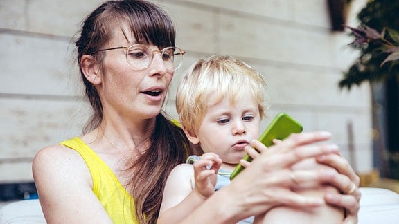 Ein kleiner Junge sitzt bei seiner Mutter auf dem Schoß und schaut auf deas Smartphone in ihrer Hand.