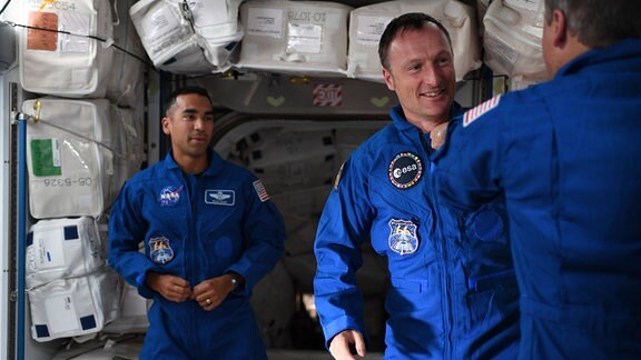NASA-Astronaut Tom Marshburn steckte ESA-Astronaut Matthias Maurer eine Anstecknadel ans Revers, die ihren neuen Status als geflogene Astronauten symbolisiert. Mashburn zeigt uns seinen Rücken am rechten Bildrand. Maurer lächelt in der Schwerelosigkeit. Im linken Bildhintergrund schaut der NASA-Astronaut  Raja Chari zu. Sie befinden sich im Inneren der Internationalen Raumstation ISS. 