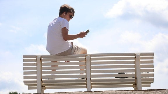 Ein Junger Mann sitzt auf der Lehne einer Parkbank und schaut auf sein Mobiltelefon.