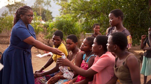 Eine Frau zeigt einer Gruppe von Schülerinnen in einem Außenbereich Menstruationsartikel 