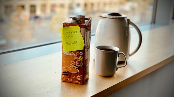 Auf einer Fensterbank stehen Kaffeekanne, eine gefüllte Tasse Kaffee und eine Packung Soja-Hafer-Drink mit Klebezettel "Schmeckt noch. Bussi", Hintergrund unscharfes Gebäude außerhalb.