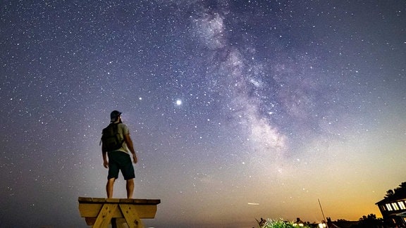 Mann steht auf einer Bank und Blickt in Sternenhimmel mit Milchstraße, Haus in der Ecke strahlt Licht aus