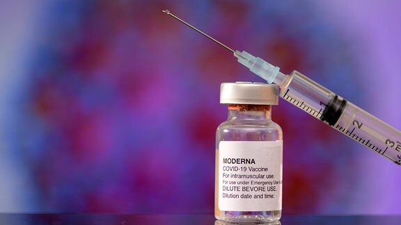 Ein Imfpstofffläschchen und eine Impfstpritze vor dem Hintergrund einer Grafik, die das Coronavirus eingefärbt in violett zeigt.