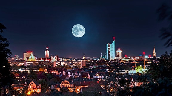 Skyline von Leipzig bei Nacht mit vielen beleuchteten Häusern und einigen Hochhäusern. Darüber Vollmond als Fotomontage.