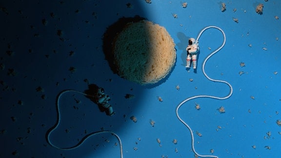 Weltraumszenerie nachgestellt mit rundem Schwamm als Mond, Krümeln als Sterne, zwei Raumfahrer*innen-Figuren mit Seilen als Schläuche, alles auf blauem Untergrund liegend.
