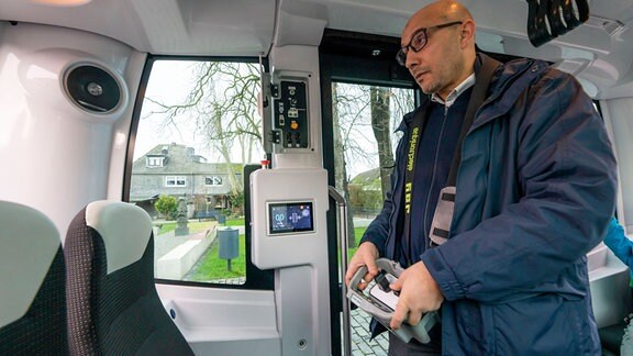Ein Mann mit Brille steht in einem Bus und hält ein Gerät mit einem Joystick in der Hand. Mit dem Joystick lenkt er den Bus.