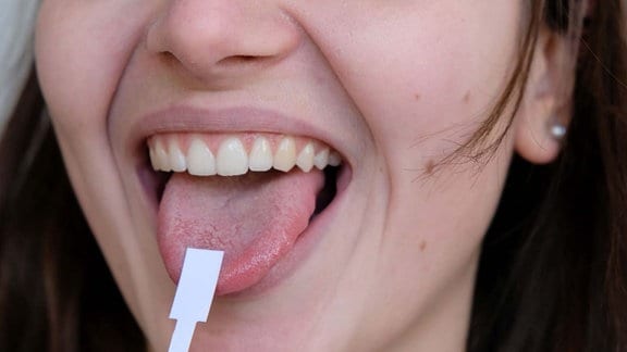 Großaufnahme der unteren Gesichtspartie einer Frau, die ihren Mund geöffnet hält und die Zunge herausstreckt. Auf der Zunge liegt ein Papierstreifen. 