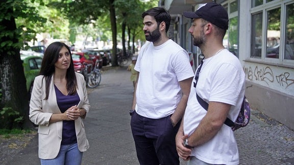 eine Junge Frau spricht mit zweri Männern in zweißen T-Shirts. Sie stehen in einer Straße auf dem Bürgersteig.