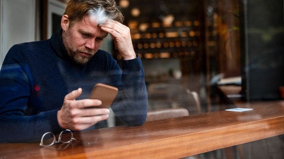 Mann mit lockigem Haare sitzt an Fenster-Tresen eines Cafés, schaut auf Smartphone und greift sich sorgenvoll am Kopf. Daneben liegt dratige Brille. Ansicht von außen durch Scheibe. Leichte unscharge Spiegelungen in Scheibe, unscharfer Hintergrund.