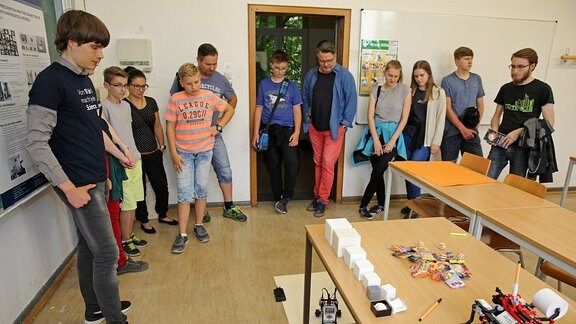 Eine Gruppe von Menschen steht im Kreis, am Fußboden liegt ein großer Bogen Papier, darauf fährt ein Roboter, der wie ein kleines Spielzeugauto aussieht, mit einem Stift über die weiße Fläche.