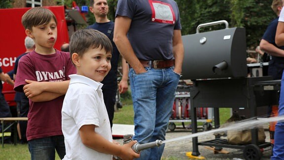 Ein junge hält eine Wasserspritze, im Hintergrund ist ein Feurwehrfahrzeug und ein Grill zu sehen.