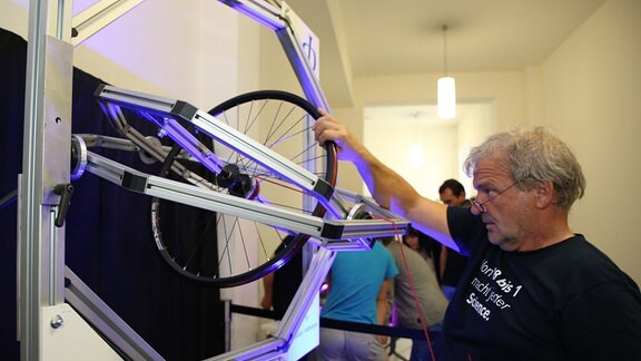 Ein Forscher steht vor einer Fahrradfelde, um die eine Schnur gewickelt ist. Durch das abziehen der Schnur lässt sich das aufgehängte Rad in Bewegung versetzen. Es ist in zwei weitere Rähmen aufgehängt, die das Rad letztlich in alle drei Dimensionen beweglich machen.