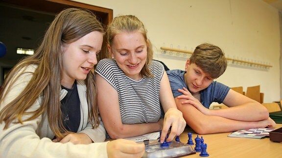 Zwei Mädchen und ein Junge spielen mit blauen Schachfiguren.