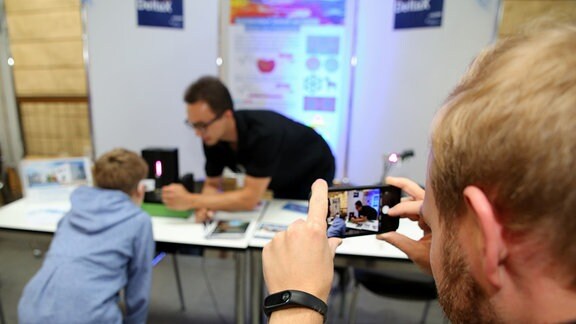 Ein Junge steht vor einem Spezialgerät, ein Forscher betätigt das Experiment. Im Vordergrund steht ein Fotograf und nimmt die Szene mit seiner Kamera auf.