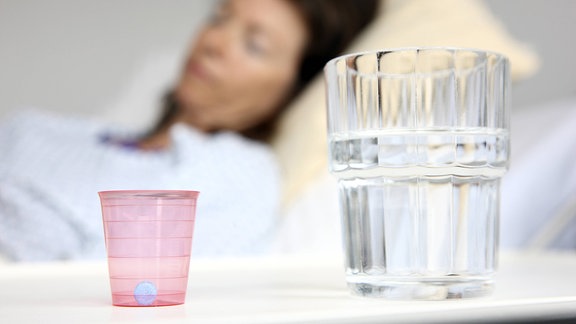 Eine Patientin in einem Krankenbett, neben ihr ein Nachttisch mit einem Wasserglas und einem Medikamentenbecher