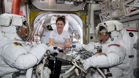 Nick Hague (mit Raumanzug), Anne McClain (mit Raumanzug) und Christina Koch in der ISS vor ihrem Weltraumspaziergang, in der ISS schwebend