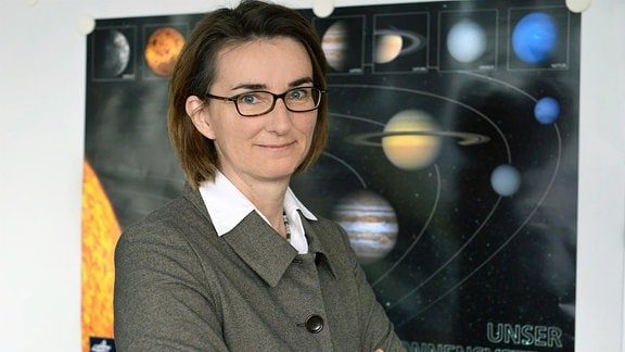 Eine Frau mit Brille, hinter ihr eine Karte des Sonnensystems.