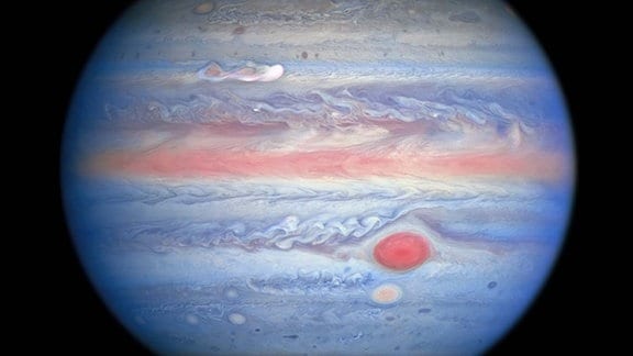 Eine vom Hubble-Weltraumteleskop der NASA / ESA am 25. August 2020 durchgeführte Multiwellenlängenbeobachtung von Jupiter im ultravioletten / sichtbaren / nahen Infrarotlicht bietet Forschern eine völlig neue Sicht auf den Riesenplaneten. Die Nahinfrarot-Bildgebung von Hubble in Kombination mit ultravioletten Ansichten bietet einen einzigartigen panchromatischen Look, der Einblicke in die Höhe und Verteilung des Dunstes und der Partikel des Planeten bietet. Dies ergänzt Hubbles Bilder mit sichtbarem Licht, die die sich ständig ändernden Wolkenmuster zeigen.  Auf diesem Foto sehen die Teile der Jupiter-Atmosphäre, die sich in größerer Höhe befinden, insbesondere über den Polen, rot aus, da atmosphärische Partikel ultraviolettes Licht absorbieren. Umgekehrt repräsentieren die blau gefärbten Bereiche das ultraviolette Licht, das vom Planeten reflektiert wird.  Ein neuer Sturm oben links, der am 18. August 2020 ausbrach, erregt die Aufmerksamkeit der Wissenschaftler in dieser Multiwellenlängenansicht. Die „Klumpen“ hinter der weißen Wolke scheinen ultraviolettes Licht zu absorbieren, ähnlich dem Zentrum des Großen Roten Flecks und des Roten Flecks Jr. direkt darunter. Dies liefert Forschern mehr Beweise dafür, dass dieser Sturm auf Jupiter länger andauern kann als die meisten Stürme.