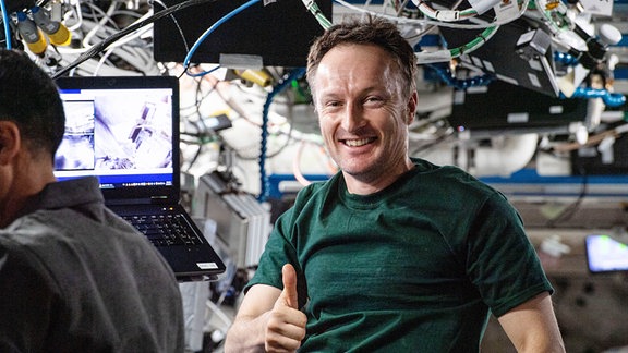 Mit einem Lächeln und hochgestrecktem Daumen blick der deutsche Esa-Astronaut Matthias Maurer in die Kamera. Er befindet sich auf der Internationalen Raumstation ISS. Im Hintergrund befinden sich Kabel an der Decke und ein aufgeklappter Laptop.